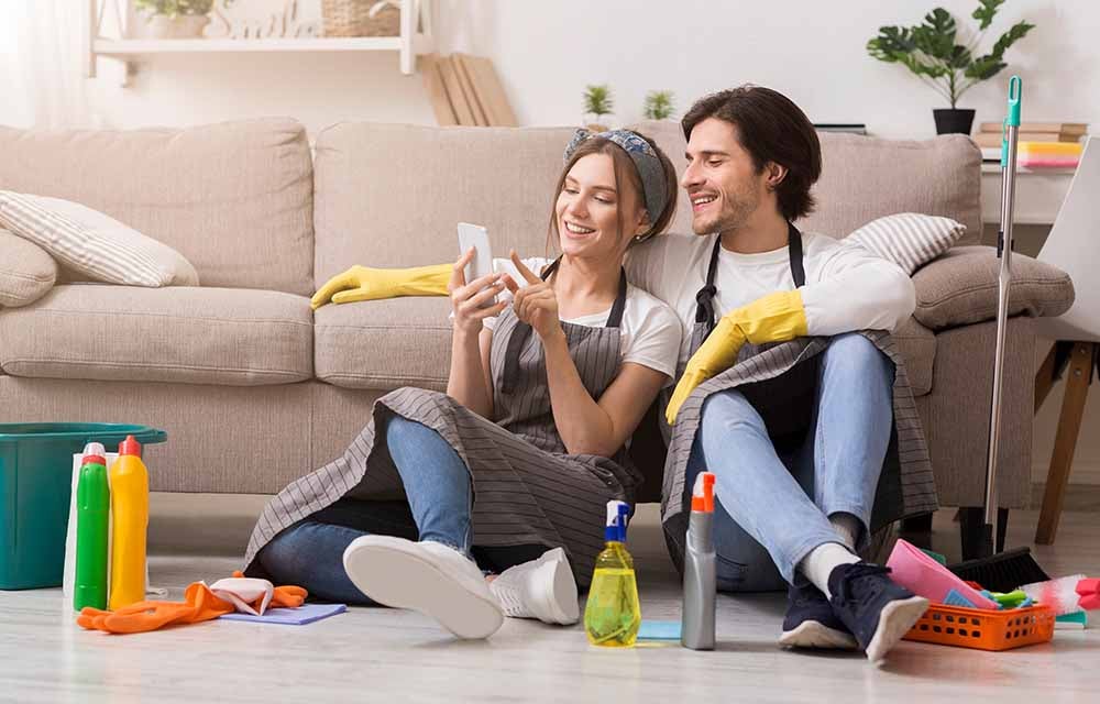 Smiling young couple relaxing with smartphone after spring-clean apartment, sitting on floor with lots of detergents and cleaning supplies, free space