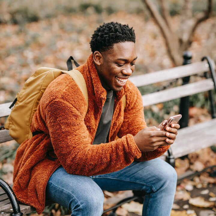 Smiling man with backpack sitting outside on a park bench looking at his phone