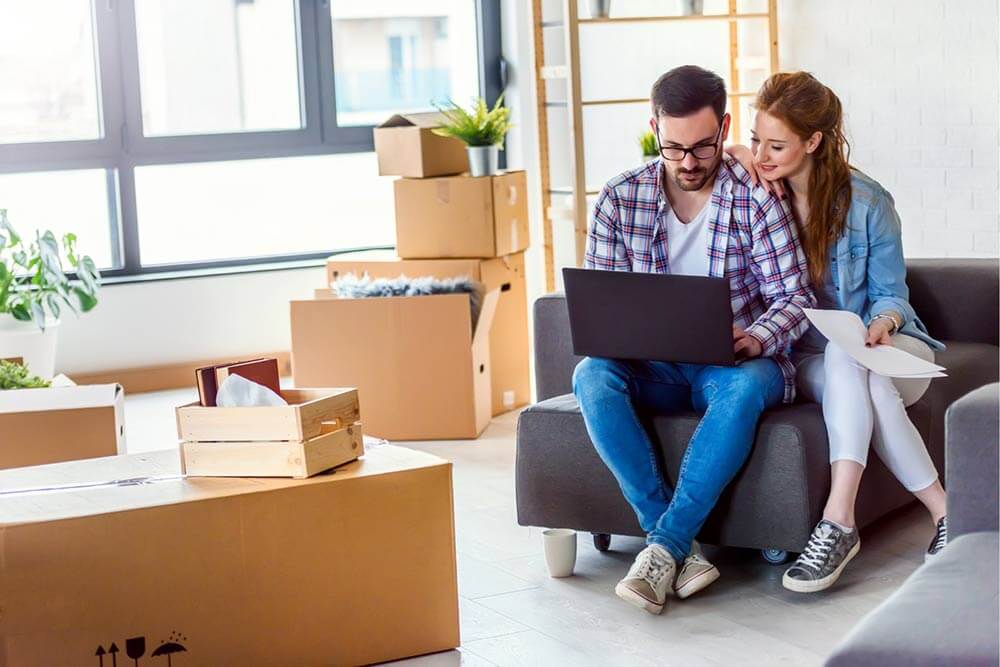 Un jeune couple emménageant dans une nouvelle maison. Assis à se détendre après avoir défait les boîtes. Effectuant des recherches sur l'assurance habitation avec un ordinateur portable.