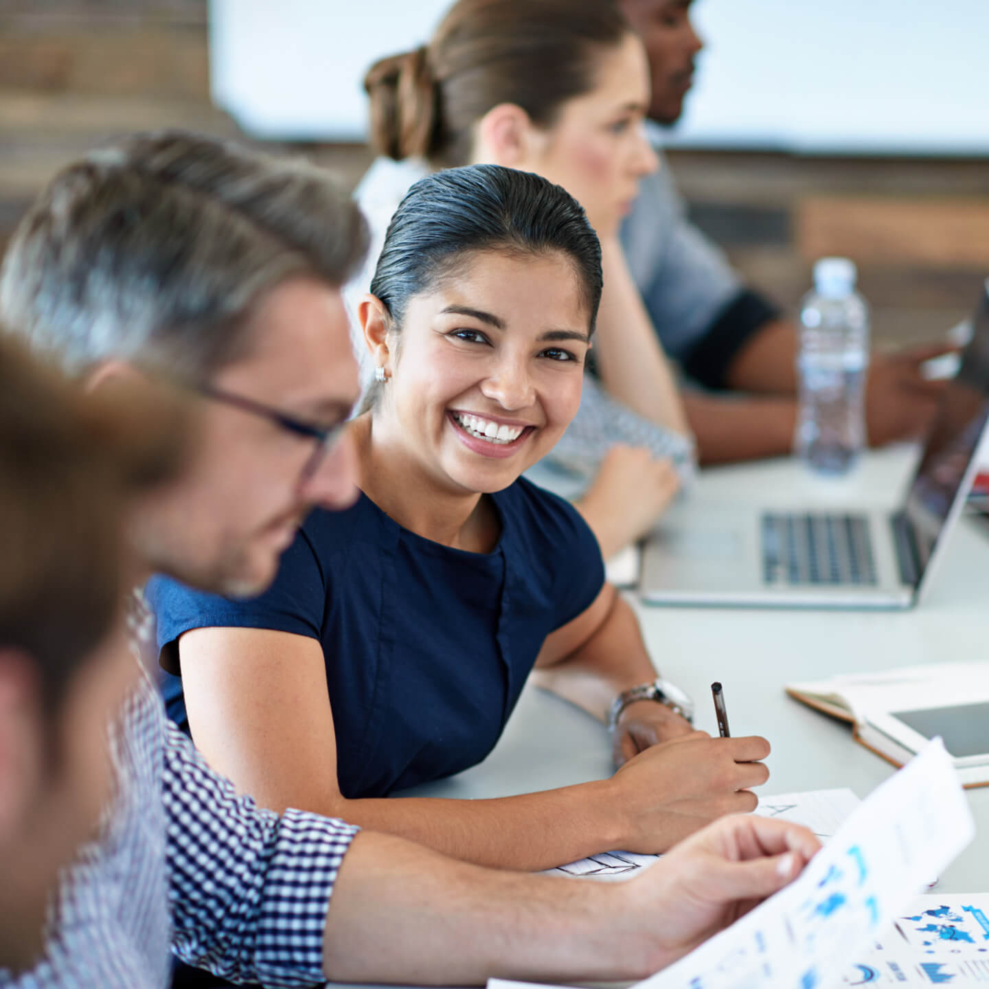A smiling woman sitting in an office meeting. She is holding a pen and is surrounded by blurry colleagues and a laptop computer.