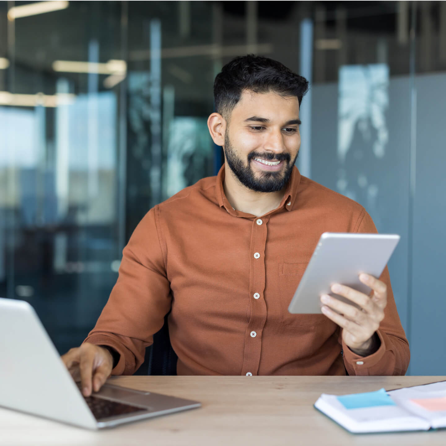 Un homme souriant, vêtu d’une chemise orangée, regarde la tablette qu’il tient dans sa main en utilisant un ordinateur portable.