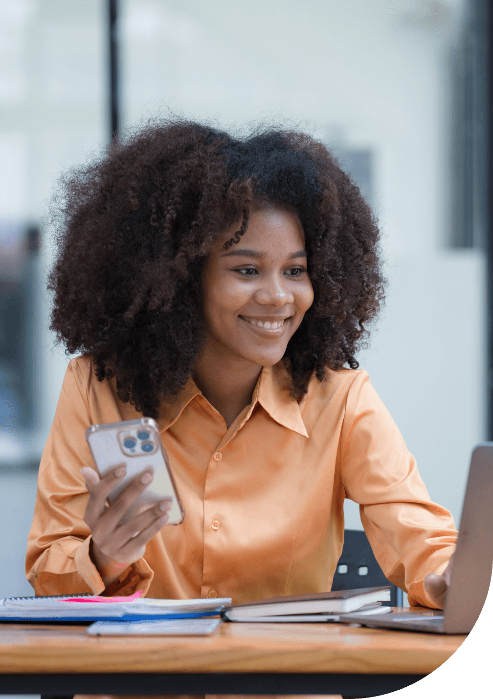 A smiling woman in an orange shirt is holding a smartphone in her hand and using a laptop computer.