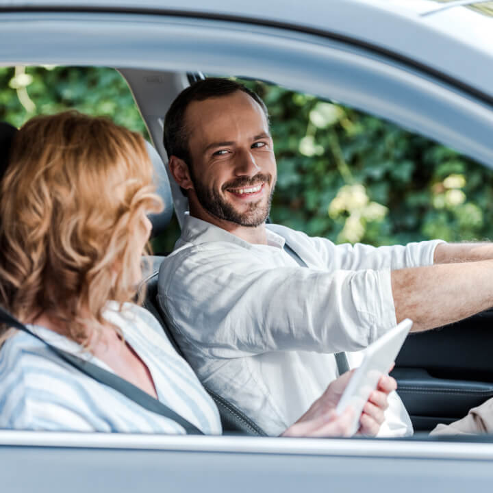 Un homme heureux conduit une voiture et observe sa femme qui utilise une tablette électronique pour trouver des directions sur les routes du Canada.