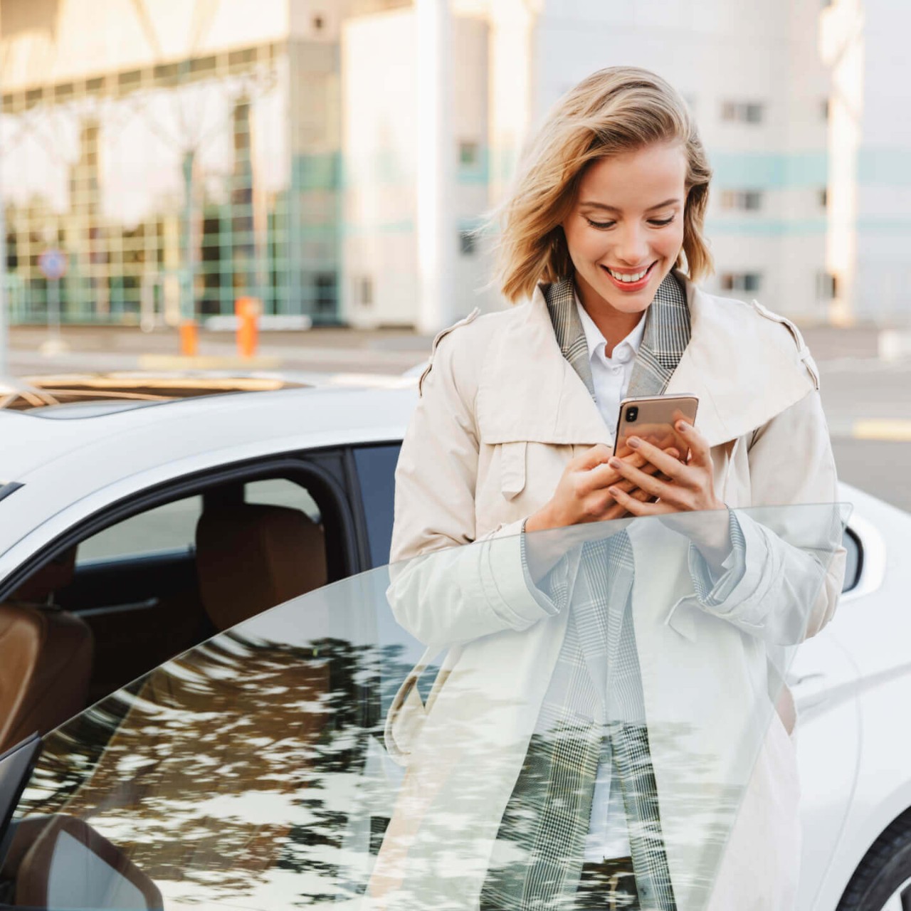 A smiling woman checking her phone while standing next to her car on a street in Toronto.