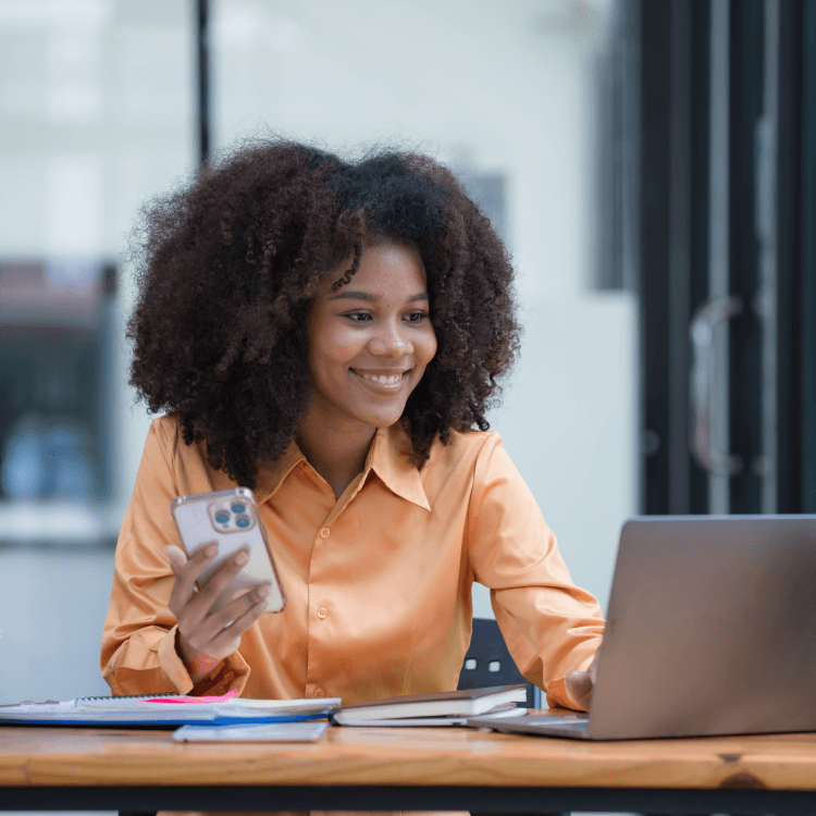 Une femme souriante, vêtue d’une chemise orangée, tient un téléphone et utilise un ordinateur portable.
