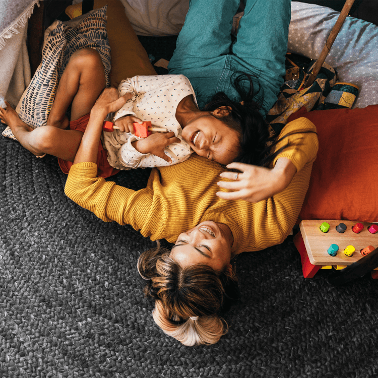 A mother and her child lying on a rug, laughing together during playtime at home.