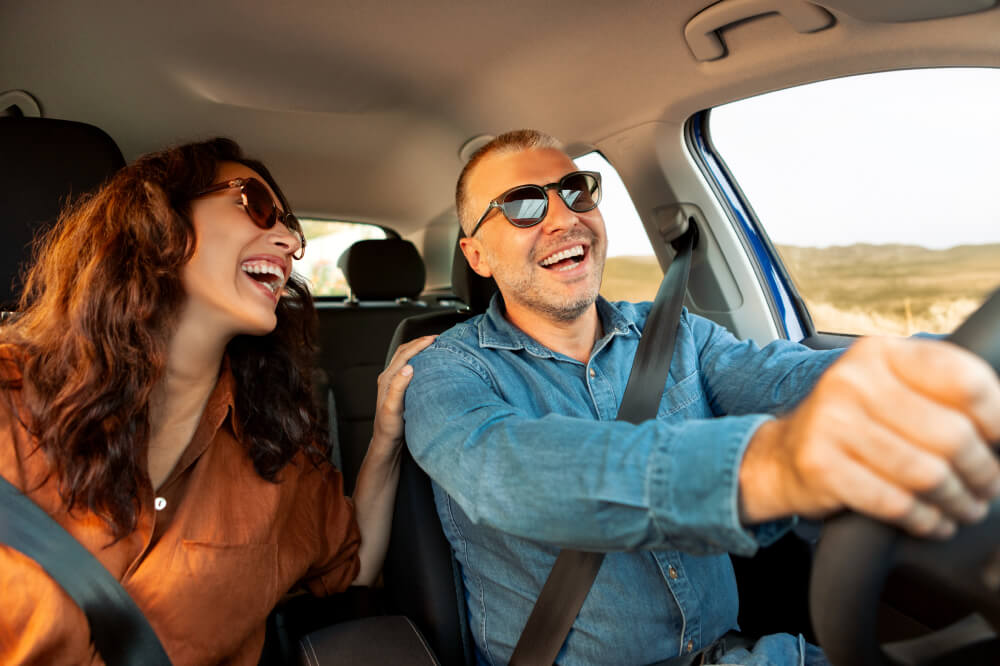 A happy couple enjoys a drive with their sunglasses on. The man smiles, looking at the road while the woman smiles back with her hand on his shoulder.
